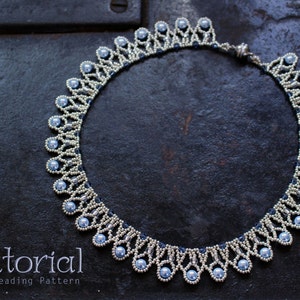 Puede incluir: Un collar de cuentas plateadas y azul claro con un diseño delicado e intrincado. El collar está hecho con cuentas pequeñas y redondas y tiene un borde festoneado. El collar está colocado sobre una superficie oscura y texturizada.  Tutorial PDF Patrón de abalorios.