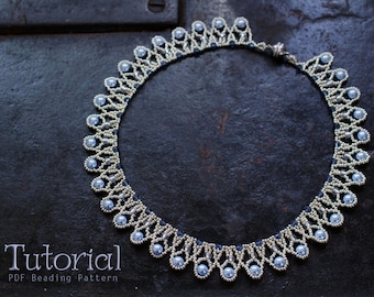 May include: A silver and light blue beaded necklace with a delicate, intricate design. The necklace is made with small, round beads and has a scalloped edge. The necklace is laid out on a dark, textured surface.  Tutorial PDF Beading Pattern.