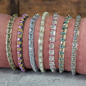 Peut inclure: Sept bracelets de perles de différentes couleurs sont présentés sur une surface rose. Les bracelets présentent une variété de perles, notamment violettes, dorées, bleues et transparentes. Chaque bracelet a un design et une palette de couleurs différents.