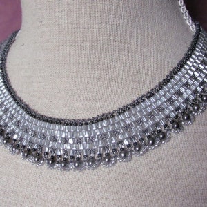 May include: A silver and gray beaded choker necklace with a graduated design. The necklace is made with small, round beads and larger, rectangular beads.