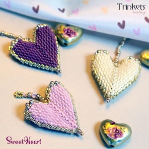 Könnte beinhalten: Nahaufnahme von herzförmigen Perlenanhängern in Lila, Rosa und Weiß mit goldfarbenem Besatz. Die Anhänger sind mit herzförmigen Charms angeordnet. Der Text "Sweet Heart" steht unten und "Trinkets beading" oben.
