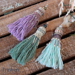 Può includere: Tre nappine fatte a mano in lavanda, verde acqua e azzurro chiaro con cime perlate. Le nappine sono etichettate "Shabby Chic tassel" e "Trinkets".