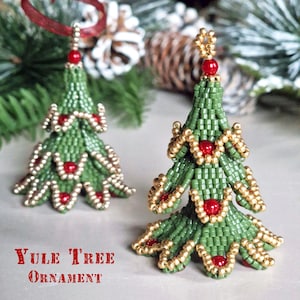 Op de afbeelding: Twee kerstboomversieringen van kralen. De bomen zijn groen met gouden accenten en rode kralenversieringen. De tekst "Yule Tree Ornament" staat in het rood onderaan de afbeelding. Eén boom heeft een rood lint om op te hangen.