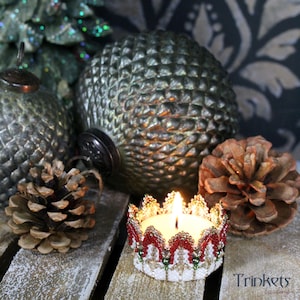 Könnte beinhalten: Ein weißer, perlenbesetzter Kerzenhalter in Form einer Krone mit roten und grünen Akzenten. Der Kerzenhalter ist angezündet und von Tannenzapfen und grünen Glaskugeln umgeben.