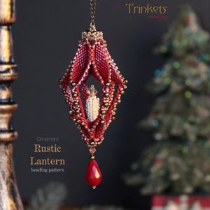Puede incluir: Un adorno de cuentas rojas y doradas con forma de linterna con una vela de cuentas blancas en su interior. El adorno está colgado de una cadena dorada y tiene una cuenta roja en forma de lágrima en la parte inferior. El adorno está etiquetado como "Linterna rústica" y "patrón de abalorios".
