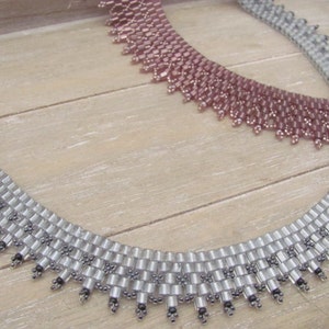 May include: Two beaded necklaces, one silver and one pink, with a scalloped edge. The necklaces are on a wooden surface.