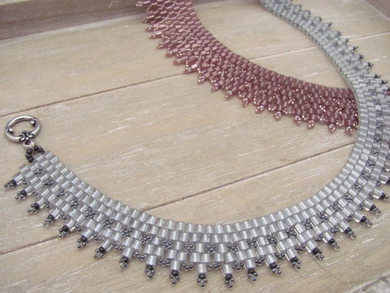 May include: Two beaded necklaces, one silver and one pink, with a scalloped edge. The necklaces are on a wooden surface.