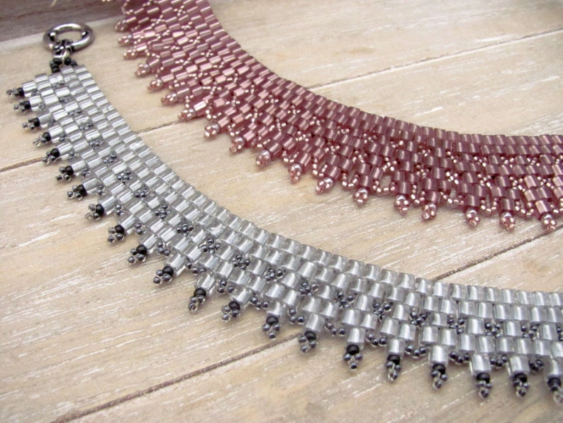 May include: Two beaded necklaces, one silver and one pink, with a fringe detail. The necklaces are made of small, square beads and are arranged in a graduated pattern.