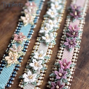 Op de afbeelding: Close-up van drie armbanden met bloemmotieven. De armbanden hebben verschillende kleuren, waaronder lichtblauw, wit en roze, met ingewikkeld kralenwerk. Het woord "Trinkets" is zichtbaar in de linkerbovenhoek.