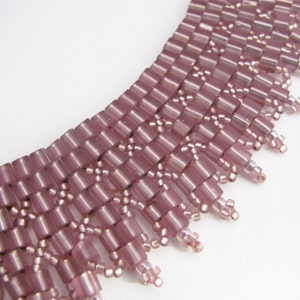 May include: A close-up of a pink beaded necklace with a fringe design. The necklace is made of small, rectangular beads that are arranged in a geometric pattern. The fringe is made of smaller beads that hang down from the bottom of the necklace.