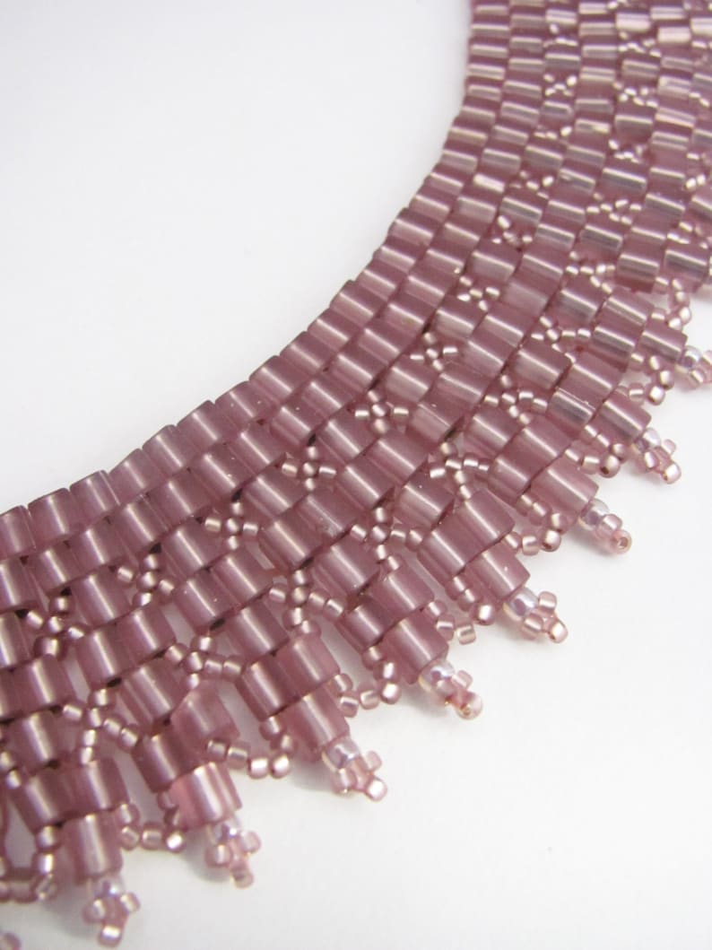 May include: A close-up of a pink beaded necklace with a fringe design. The necklace is made of small, rectangular beads that are arranged in a geometric pattern. The fringe is made of smaller beads that hang down from the bottom of the necklace.