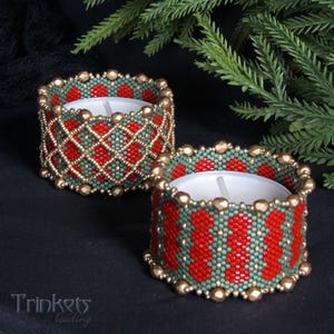 Puede incluir: Dos portavelas de cuentas rojas, verdes y doradas. Los portavelas están hechos con cuentas pequeñas y tienen un patrón geométrico. Los portavelas están sobre una superficie negra.