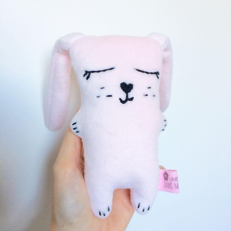May include: A soft, pink plush bunny with black embroidered facial features, including closed eyes, a heart-shaped nose, and whiskers. The bunny has long ears and small paws. A pink tag with white text is attached to the bunny.