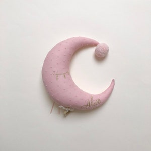 K&ouml;nnte beinhalten: Ein rosa halbmondf&ouml;rmiges Kissen mit einem goldenen gestickten Auge und dem Namen "Alice" auf der Unterseite. Das Kissen hat einen kleinen rosa Bommel oben.