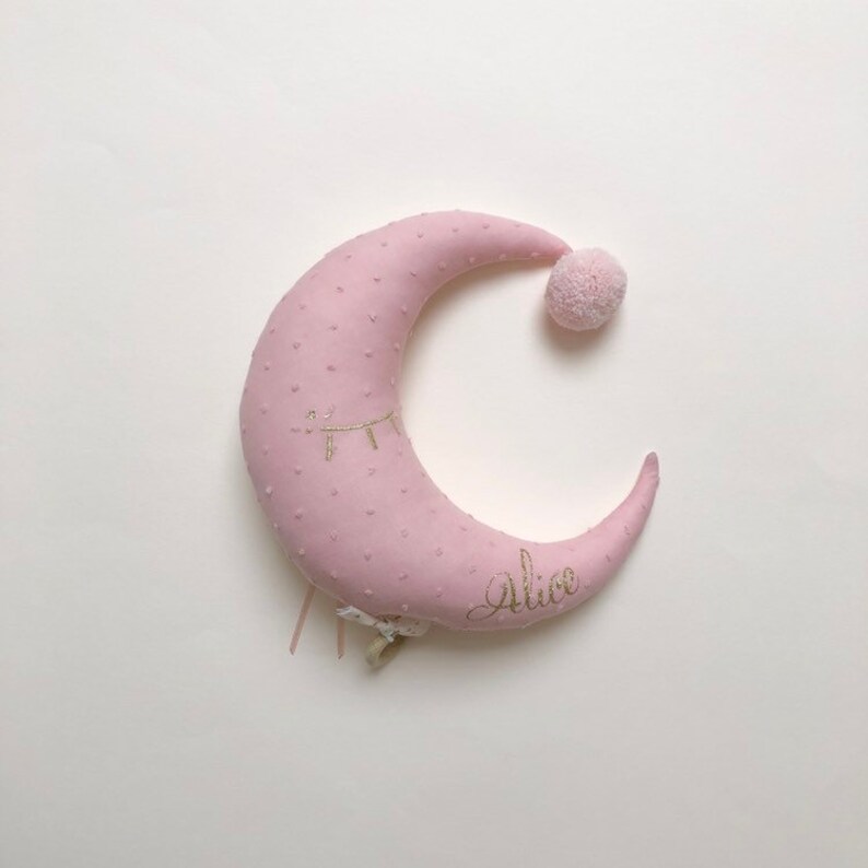 K&ouml;nnte beinhalten: Ein rosa halbmondf&ouml;rmiges Kissen mit einem goldenen gestickten Auge und dem Namen "Alice" auf der Unterseite. Das Kissen hat einen kleinen rosa Bommel oben.