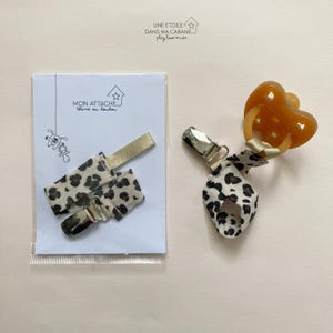 May include: A beige and brown leopard print pacifier clip with a silver clip. The clip is attached to a pacifier with a brown silicone ring. The clip is packaged in a clear plastic bag with a white card that says "Mon Attache" and "tetine ou doudou".