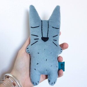 May include: A light blue felt cat doll with black embroidered facial features and small black spots. The cat has pointed ears and a small, dark blue tag attached to its side. The doll is approximately 6 inches tall.