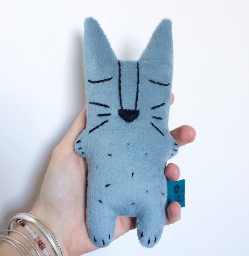 May include: A light blue felt cat doll with black embroidered facial features and small black spots. The cat has pointed ears and a small, dark blue tag attached to its side. The doll is approximately 6 inches tall.
