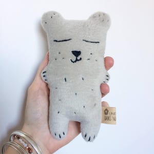 May include: A handmade, light gray plush bear with black embroidered details. The bear has closed eyes, a small black nose, and black spots. A small tag is attached to the bear's side. The bear is held in a hand.