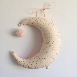 K&ouml;nnte beinhalten: Ein rosa halbmondf&ouml;rmiges Kissen mit einem goldenen Dreiecksmuster. Das Kissen hat einen wei&szlig;en Bommel oben und einen wei&szlig;en Knopf unten. Das Kissen h&auml;ngt an einem rosa Band.