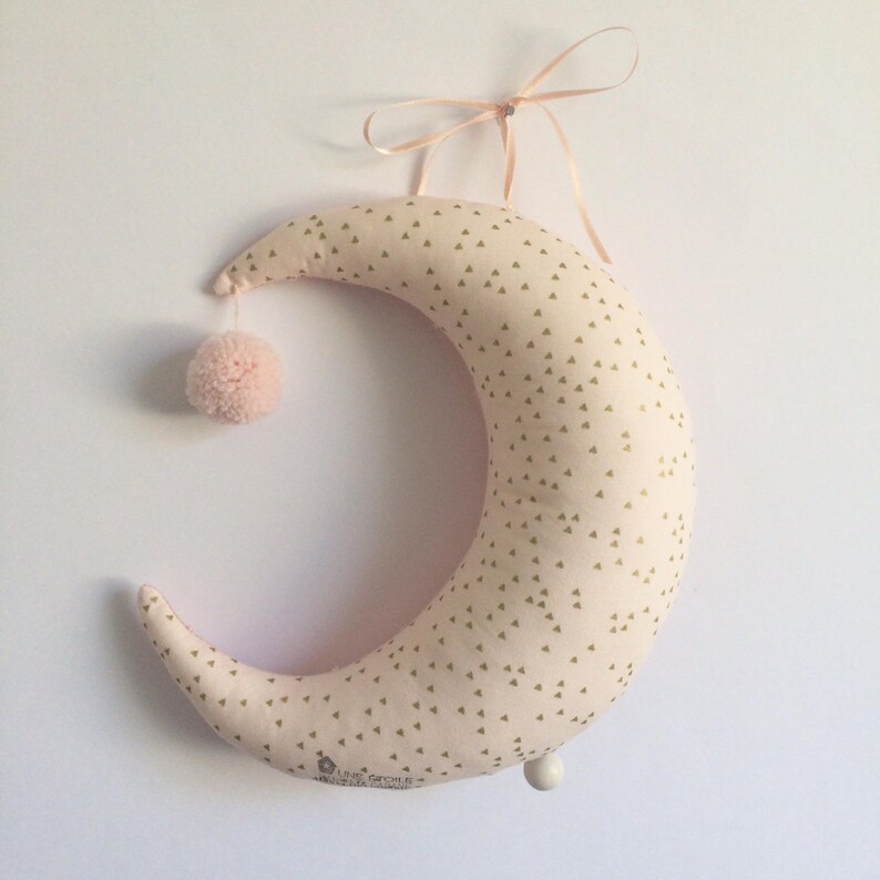 K&ouml;nnte beinhalten: Ein rosa halbmondf&ouml;rmiges Kissen mit einem goldenen Dreiecksmuster. Das Kissen hat einen wei&szlig;en Bommel oben und einen wei&szlig;en Knopf unten. Das Kissen h&auml;ngt an einem rosa Band.