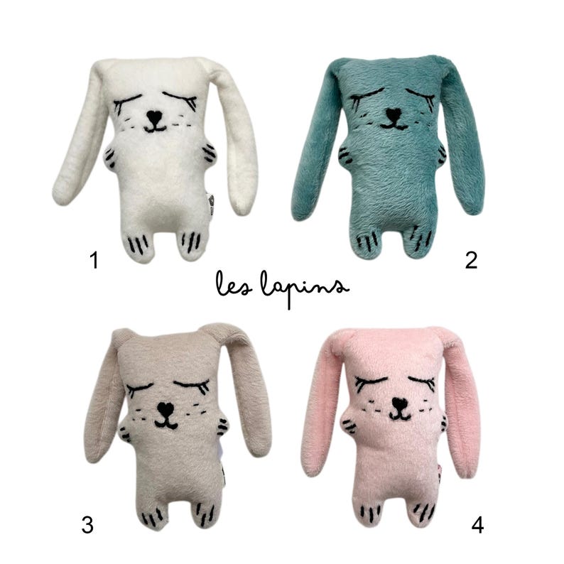 May include: Four plush bunny toys in white, teal, tan, and pink. Each bunny has long ears, closed eyes, and a black heart-shaped nose. The text "les lapins" is written in black script below the bunnies.