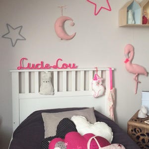 K&ouml;nnte beinhalten: Ein wei&szlig;es Bett mit einem grauen und rosafarbenen Bettbezug. Auf dem Bett liegen mehrere Kissen, darunter ein rosafarbenes, wolkenf&ouml;rmiges Kissen mit dem Text "Lucie Lou" darauf. Die Wand hinter dem Bett ist wei&szlig; und mit rosafarbenen Sternen, einem rosafarbenen Mond und einem rosafarbenen Flamingo verziert.