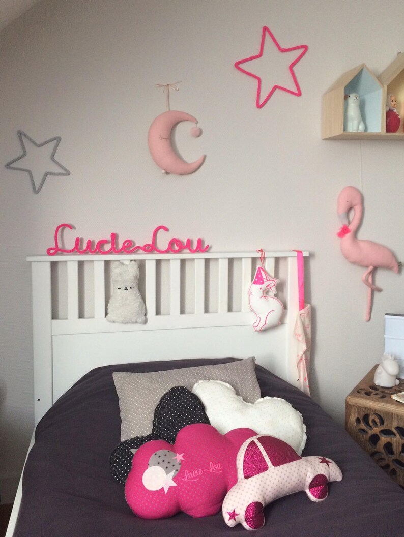 K&ouml;nnte beinhalten: Ein wei&szlig;es Bett mit einem grauen und rosafarbenen Bettbezug. Auf dem Bett liegen mehrere Kissen, darunter ein rosafarbenes, wolkenf&ouml;rmiges Kissen mit dem Text "Lucie Lou" darauf. Die Wand hinter dem Bett ist wei&szlig; und mit rosafarbenen Sternen, einem rosafarbenen Mond und einem rosafarbenen Flamingo verziert.