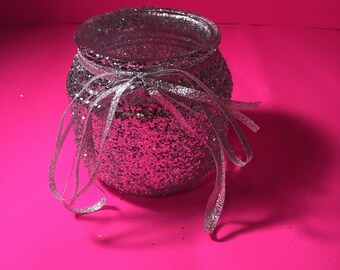 Puede incluir: Un tarro de cristal plateado con purpurina y una cinta plateada atada alrededor de la parte superior. El tarro está sobre una superficie rosa.