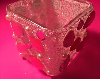 Puede incluir: Un jarrón de vidrio transparente en forma de cubo decorado con purpurina y pedrería rosa, roja y blanca. El jarrón es perfecto para exhibir flores pequeñas u otros artículos decorativos.