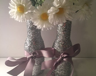 Puede incluir: Dos botellas de vidrio con purpurina plateada con flores de margarita blancas y atadas con una cinta de color morado claro.