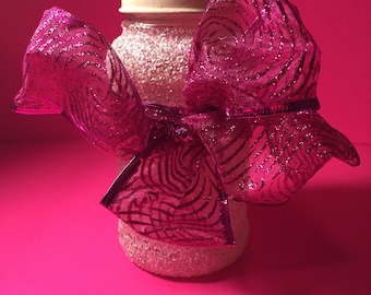 Puede incluir: Un tarro de vidrio transparente con tapa blanca, decorado con purpurina, está atado con un gran lazo rosa con un patrón brillante a rayas.