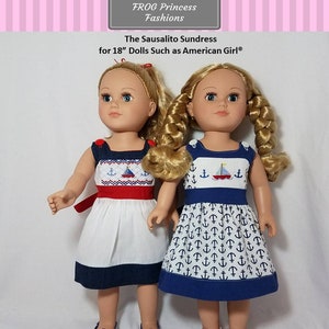 May include: Two 18-inch dolls wearing white and blue sundresses with red and blue embroidery. The dresses have a nautical theme with anchors and sailboats. The dolls have blonde hair and blue eyes. The text on the sign reads "FROG Princess Fashions The Sausalito Sundress for 18" Dolls Such as American Girl®" and "A Machine Embroidery Design Made ITH for Large Hoops Only".
