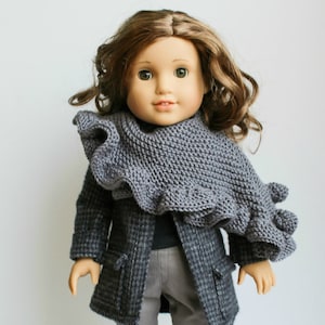 Könnte beinhalten: Ein grauer gestrickter Schal mit einem gekräuselten Rand, entworfen für 45,7 cm Puppen. Der Schal wird von einer Puppe mit braunen Haaren und einem grauen Mantel getragen. Der Text "Dana Shawl" und "A Little Knitty" ist im Bild sichtbar.