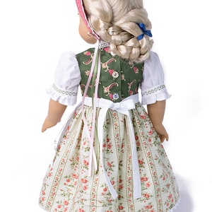 May include: A doll wearing a green and white dirndl with a pink ribbon. The dirndl has a floral pattern and a white lace trim. The doll has blonde hair and is wearing a blue ribbon in her hair.