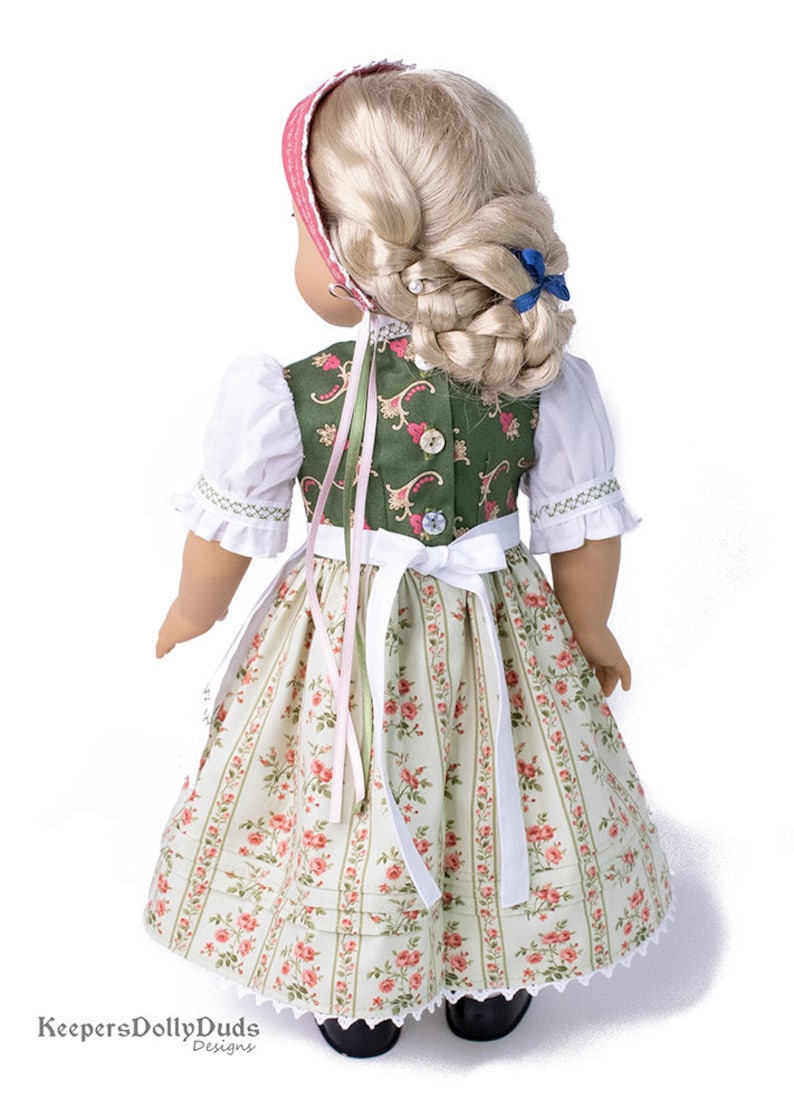 May include: A doll wearing a green and white dirndl with a pink ribbon. The dirndl has a floral pattern and a white lace trim. The doll has blonde hair and is wearing a blue ribbon in her hair.