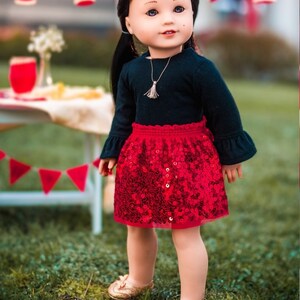 Pu&ograve; includere: Una bambola che indossa una camicia nera a maniche lunghe con una gonna rossa di paillettes. La bambola ha i capelli castani e gli occhi azzurri. La bambola &egrave; in piedi sull'erba verde con decorazioni rosse e bianche sullo sfondo.