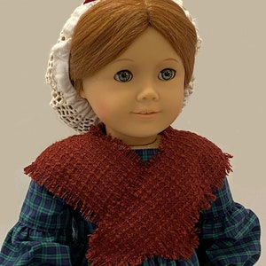 May include: A doll wearing a green and blue plaid dress with a brown shawl and a white lace bonnet with a burgundy bow.