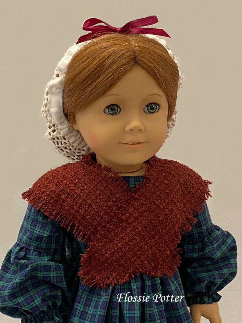 May include: A doll wearing a green and blue plaid dress with a brown shawl and a white lace bonnet with a burgundy bow.