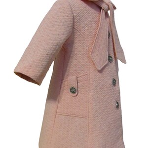 Puede incluir: Un abrigo acolchado rosa con un gran lazo de corbata y botones plateados. El abrigo tiene un bolsillo en el lado izquierdo.