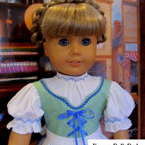 May include: A doll wearing a white blouse with blue trim and a green bodice with blue ribbon lacing. The doll has blonde hair in braids and blue eyes. The doll is wearing a white skirt with a green and white pattern. The doll is standing in front of a backdrop with a building and a sign that says "KeepersDollyDuds Designs".