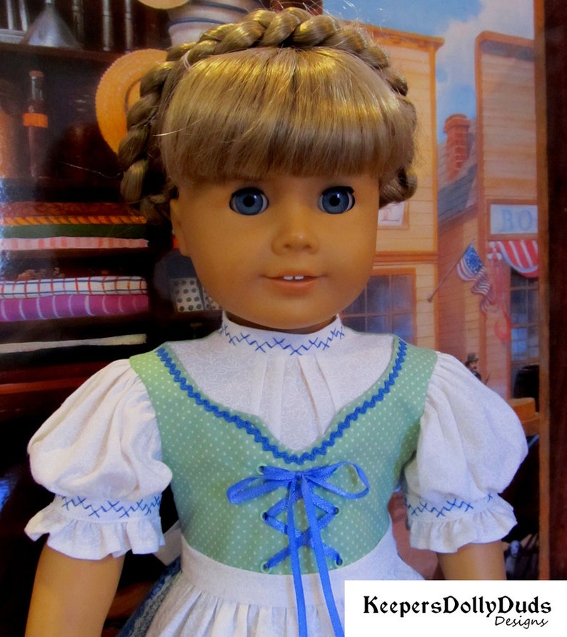 May include: A doll wearing a white blouse with blue trim and a green bodice with blue ribbon lacing. The doll has blonde hair in braids and blue eyes. The doll is wearing a white skirt with a green and white pattern. The doll is standing in front of a backdrop with a building and a sign that says "KeepersDollyDuds Designs".