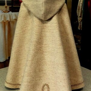 May include: A beige hooded cape with brown trim. The cape has a chain-like design around the hood and hem. The cape is made of a soft, textured fabric. Keepers Dolly Duds.