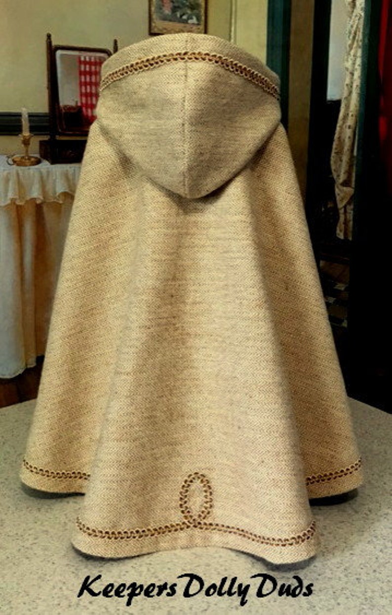 May include: A beige hooded cape with brown trim. The cape has a chain-like design around the hood and hem. The cape is made of a soft, textured fabric. Keepers Dolly Duds.