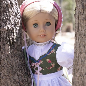 May include: A doll wearing a green and white dirndl with a pink ribbon and a white blouse. The doll is wearing a pink and white bonnet and has blonde hair and blue eyes. The doll is standing in front of a tree.