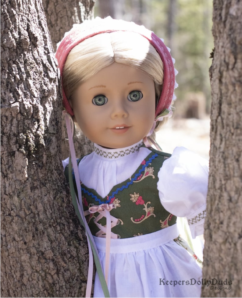 May include: A doll wearing a green and white dirndl with a pink ribbon and a white blouse. The doll is wearing a pink and white bonnet and has blonde hair and blue eyes. The doll is standing in front of a tree.