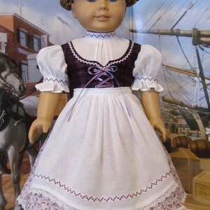 May include: A doll wearing a white dress with a burgundy bodice and a purple ribbon. The dress has a lace trim and embroidered details. The doll has blonde hair and blue eyes. The doll is wearing brown shoes. The image is captioned "KeepersDollyDuds Designs".