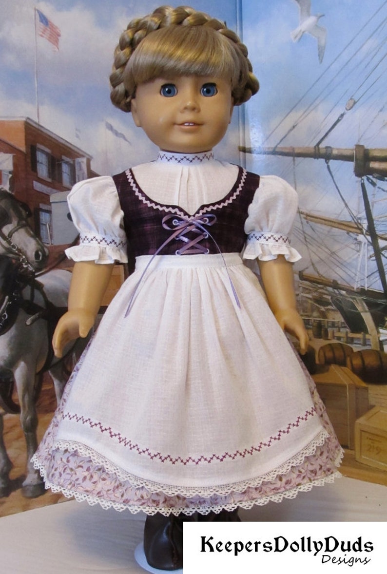 May include: A doll wearing a white dress with a burgundy bodice and a purple ribbon. The dress has a lace trim and embroidered details. The doll has blonde hair and blue eyes. The doll is wearing brown shoes. The image is captioned "KeepersDollyDuds Designs".