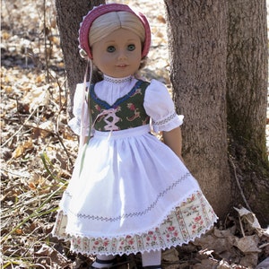 May include: A doll wearing a white dress with a green and floral bodice, a pink and white hat, and white socks and black shoes. The doll is standing in front of a tree trunk.