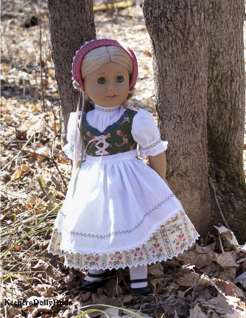 May include: A doll wearing a white dress with a green and floral bodice, a pink and white hat, and white socks and black shoes. The doll is standing in front of a tree trunk.